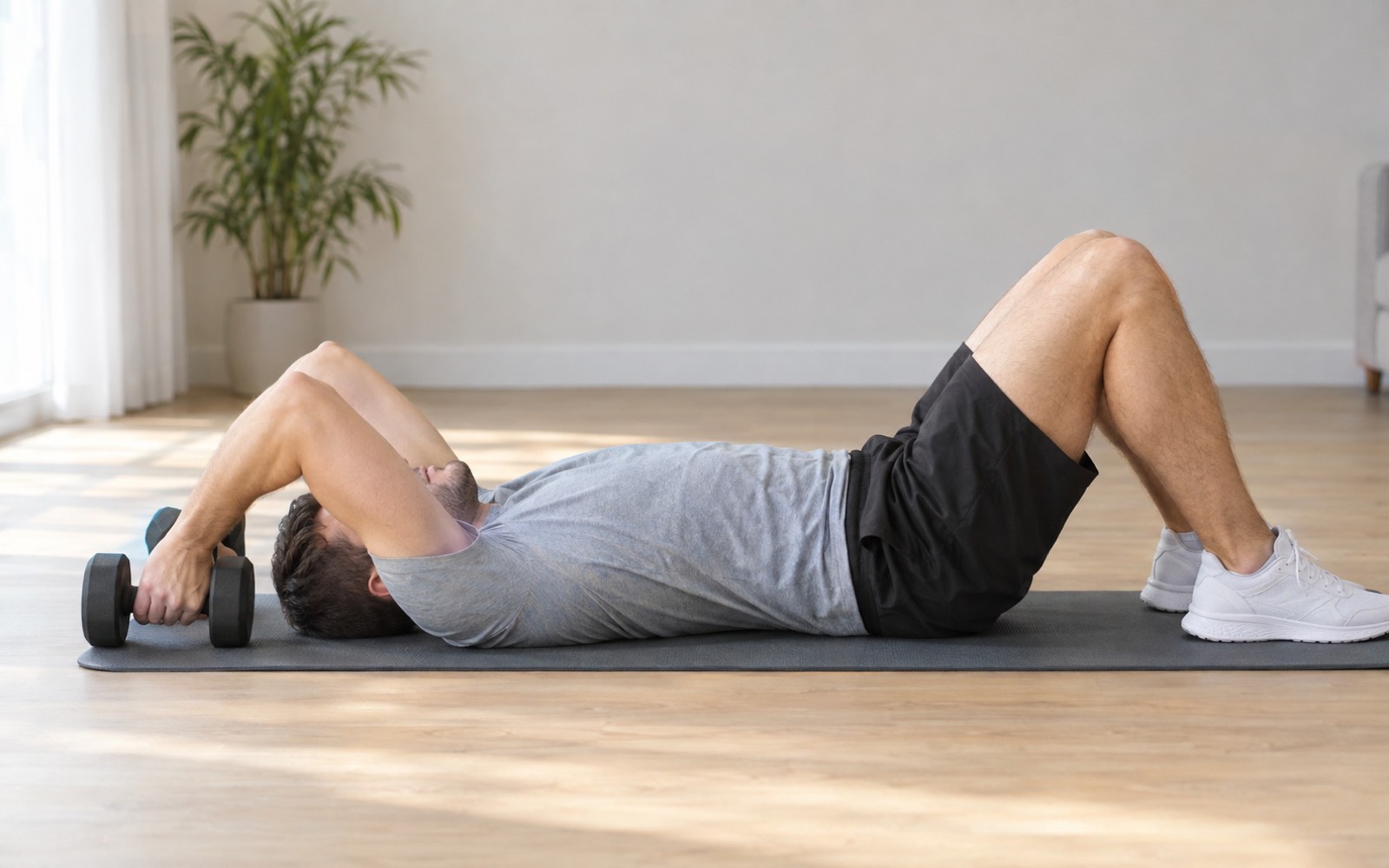 A man lying on a mat performing dumbbell skull crushers with a neutral grip, dumbbells lowered behind his head to the floor.