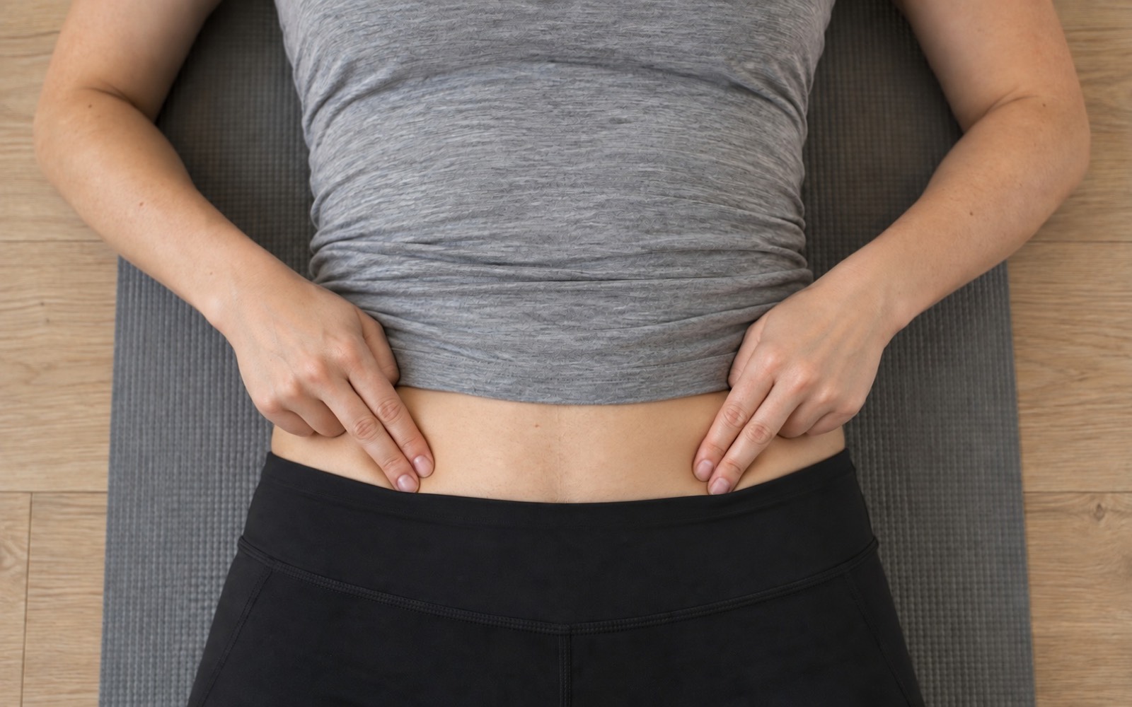 Top view: a woman lying on her back presses two fingertips of each hand into her lower belly just inside the hip bones to check her stomach vacuum technique