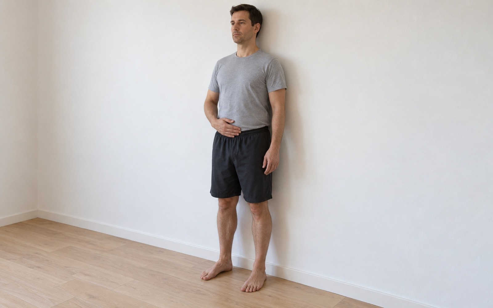 Man standing with his back to the wall, one palm on his lower belly, performing a standing stomach vacuum